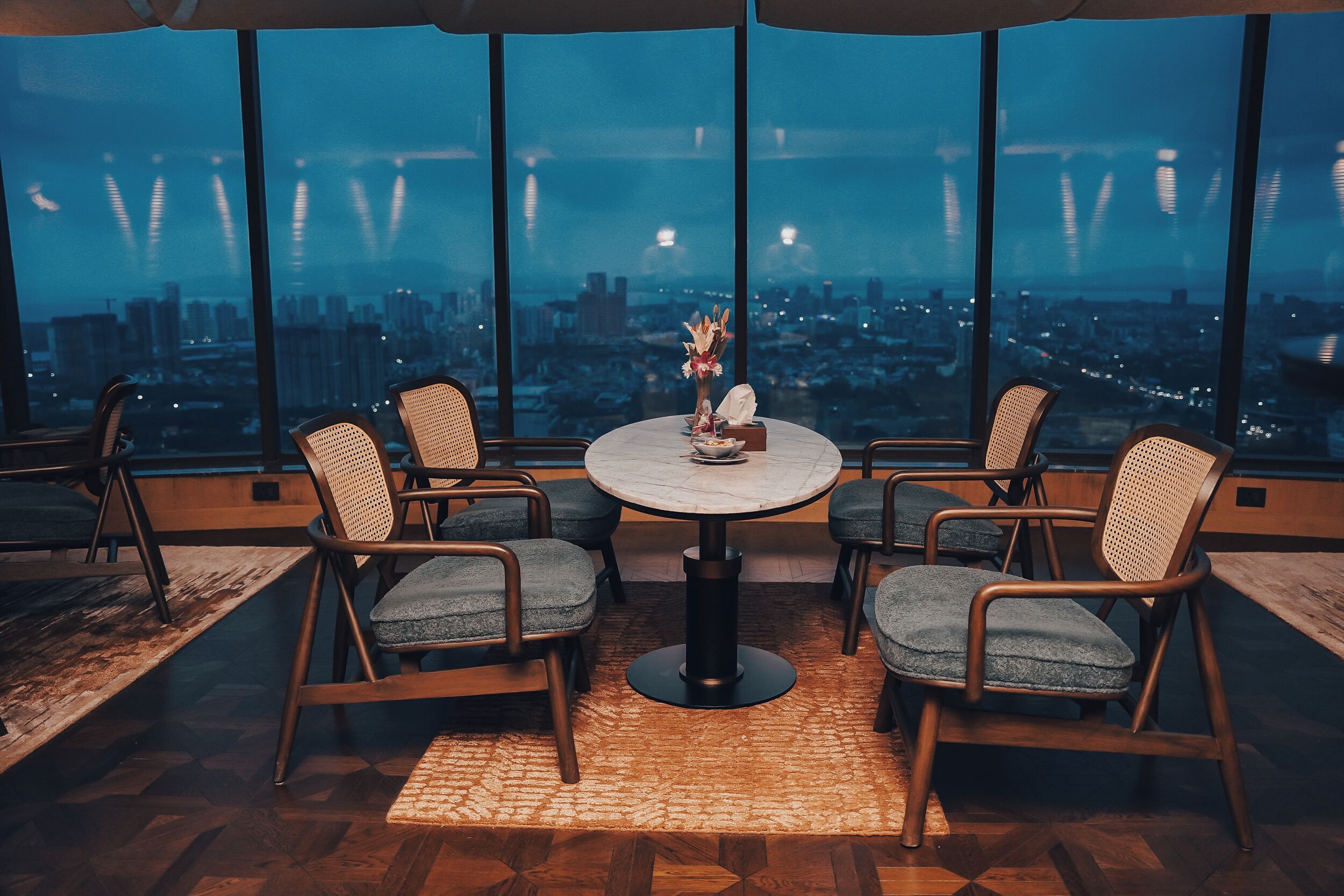 A dining area with a round table and six chairs, large windows with a city view, and a wooden floor.