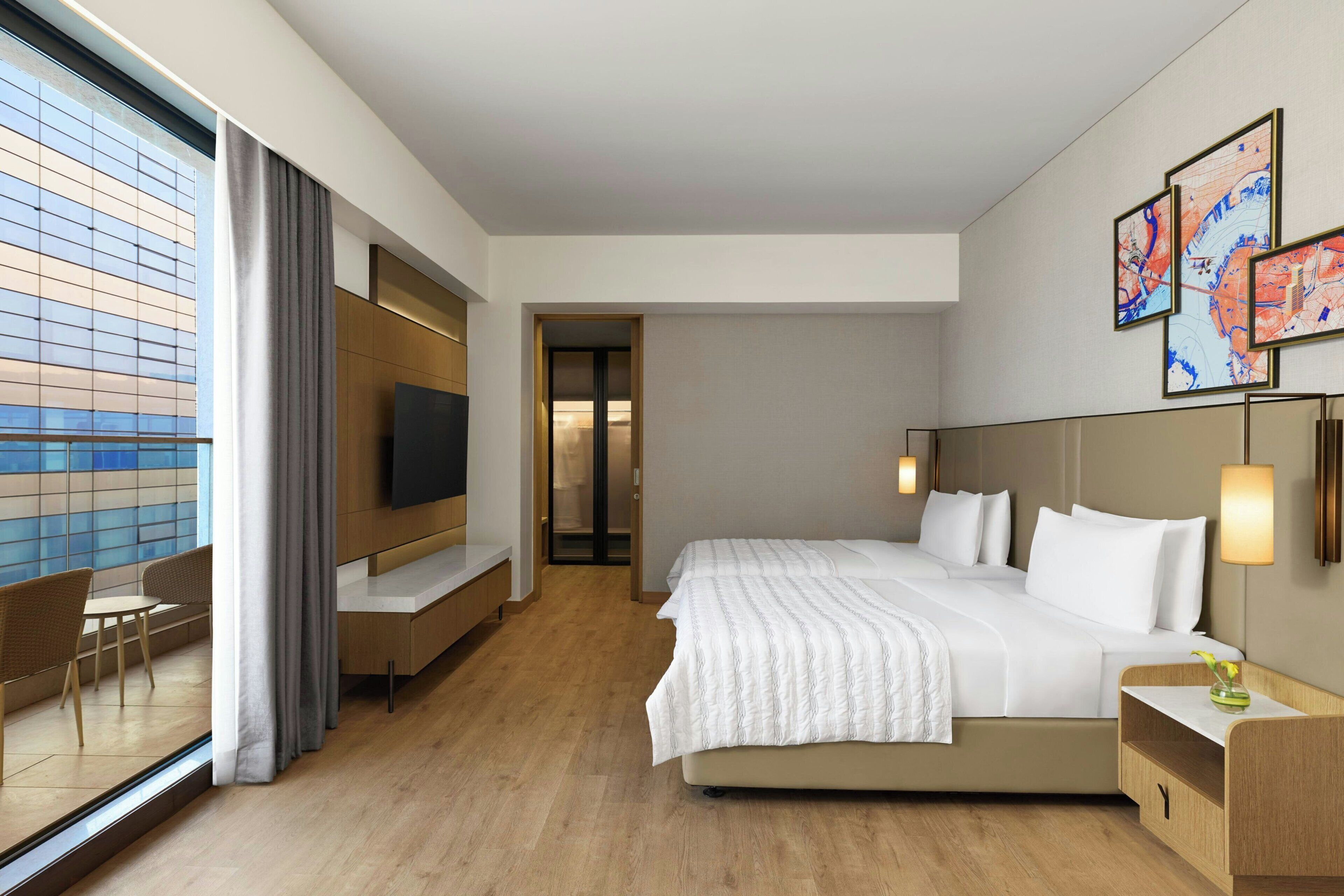 A modern hotel room with a large bed, a desk, a TV, and a view of the city through the window.