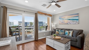 A living room with a sliding glass door to a balcony, a ceiling fan, a painting of a beach, and a grey sofa with decorative pillows. - NEW Heated Pool & HOT Tub! Top Floor! Elevator (Galveston)