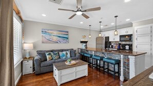 A modern kitchen with a bar area, a gray sofa with decorative pillows, a wooden coffee table, and a ceiling fan. - NEW Heated Pool & HOT Tub! Top Floor! Elevator (Galveston)