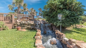 A small waterfall in a landscaped area with a wooden deck and a warning sign. - NEW Heated Pool & HOT Tub! Top Floor! Elevator (Galveston)