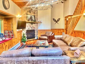 A cozy living room with a stone fireplace, leather sofas, a television, and a wooden staircase.