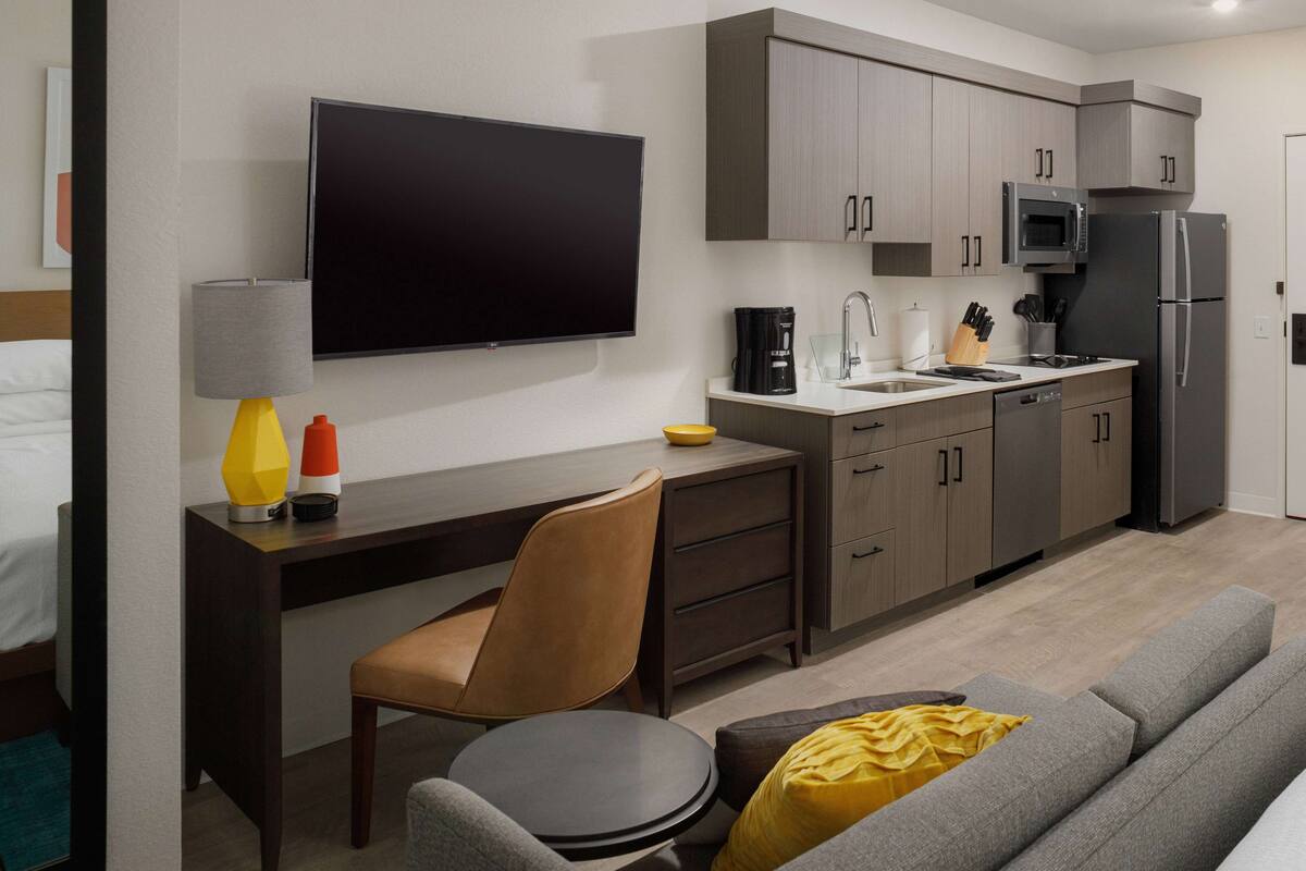 A modern hotel room with a kitchenette, a flat-screen TV, a sofa, and a dining table.
