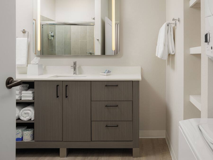 A modern bathroom with a white sink, a mirror, and a shower. There are towels and toiletries on the counter.