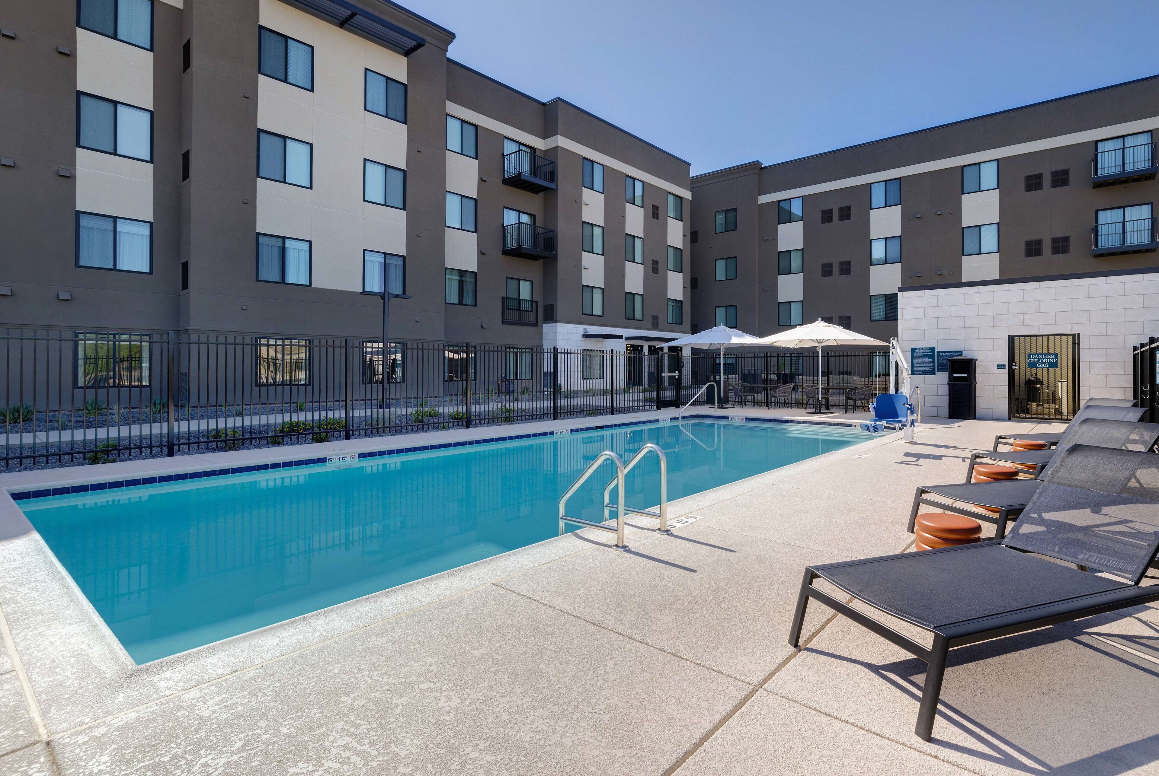 A swimming pool with lounge chairs and umbrellas, surrounded by a fence and a building with multiple balconies.