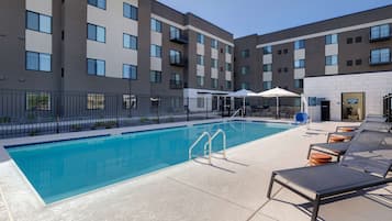 A swimming pool with lounge chairs and umbrellas, surrounded by a fence and a building with multiple balconies.