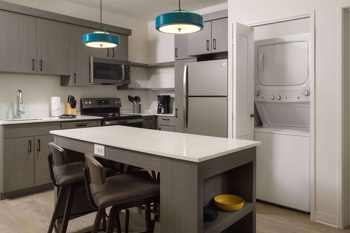A modern kitchen with grey cabinets, a central island with bar stools, a microwave, a coffee maker, and a built-in washer and dryer.
