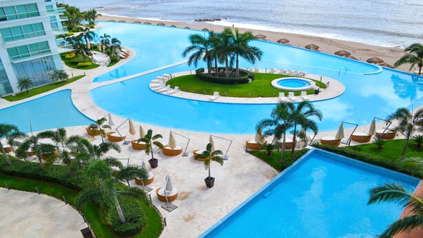 A resort with a large central pool, smaller surrounding pools, palm trees, and a beach view.