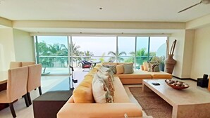 A modern living room with a large sofa, a dining area, and a view of palm trees through large windows.