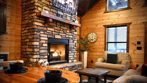 Ein gemütliches Blockhäuschen im Inneren mit Kamin aus Stein, Holzwänden, einer Couch und einem Esstisch.