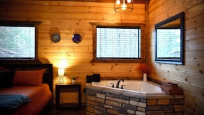Ein Holzblockhaus-Interieur mit einer steinernen Badewanne, einem Bett mit orangefarbener Bettwäsche und einem kleinen Tisch mit Lampe und Pflanze.