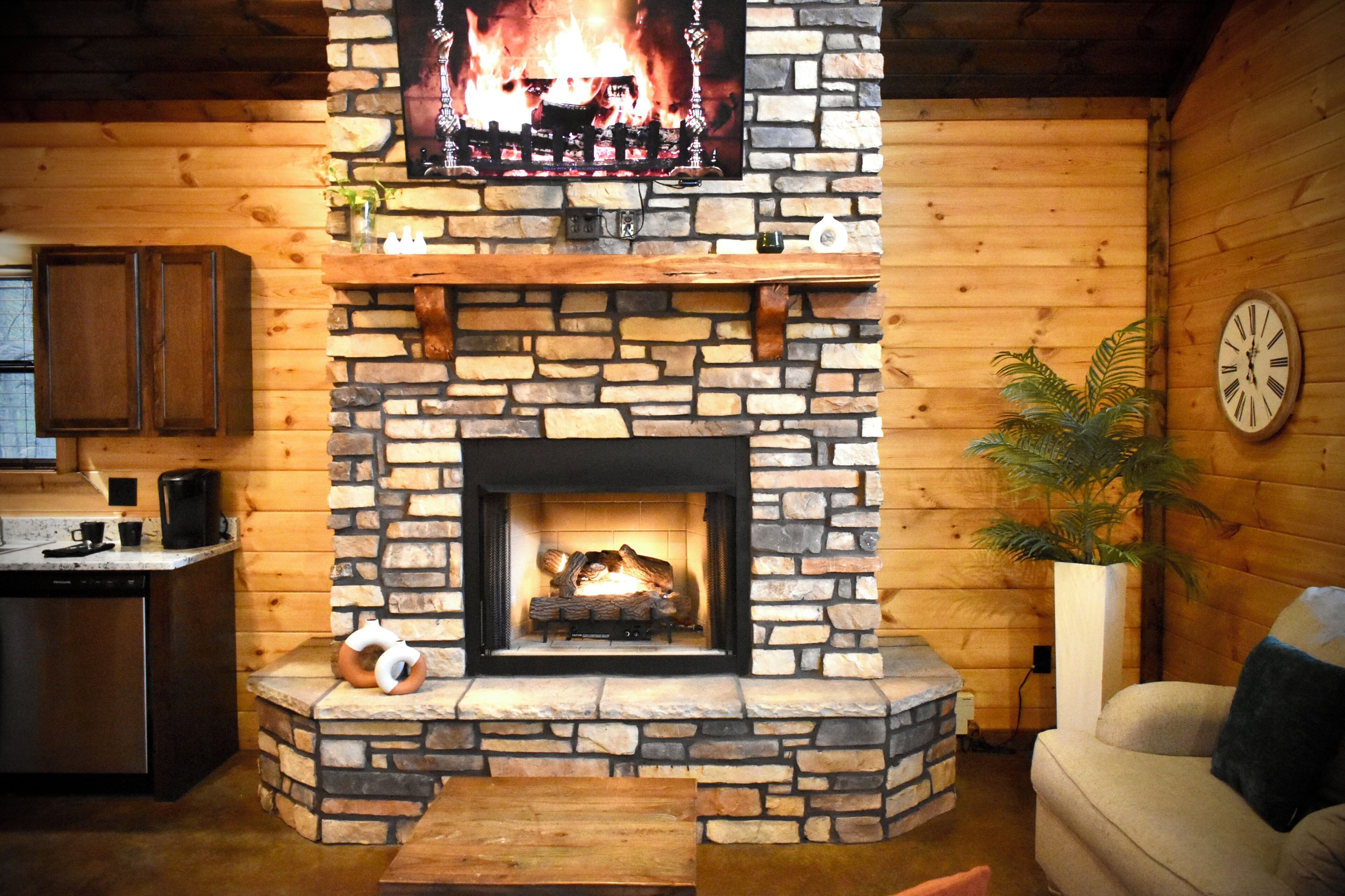 Una chimenea de piedra con un televisor encima, un banco de madera, un sofá y un reloj en la pared.