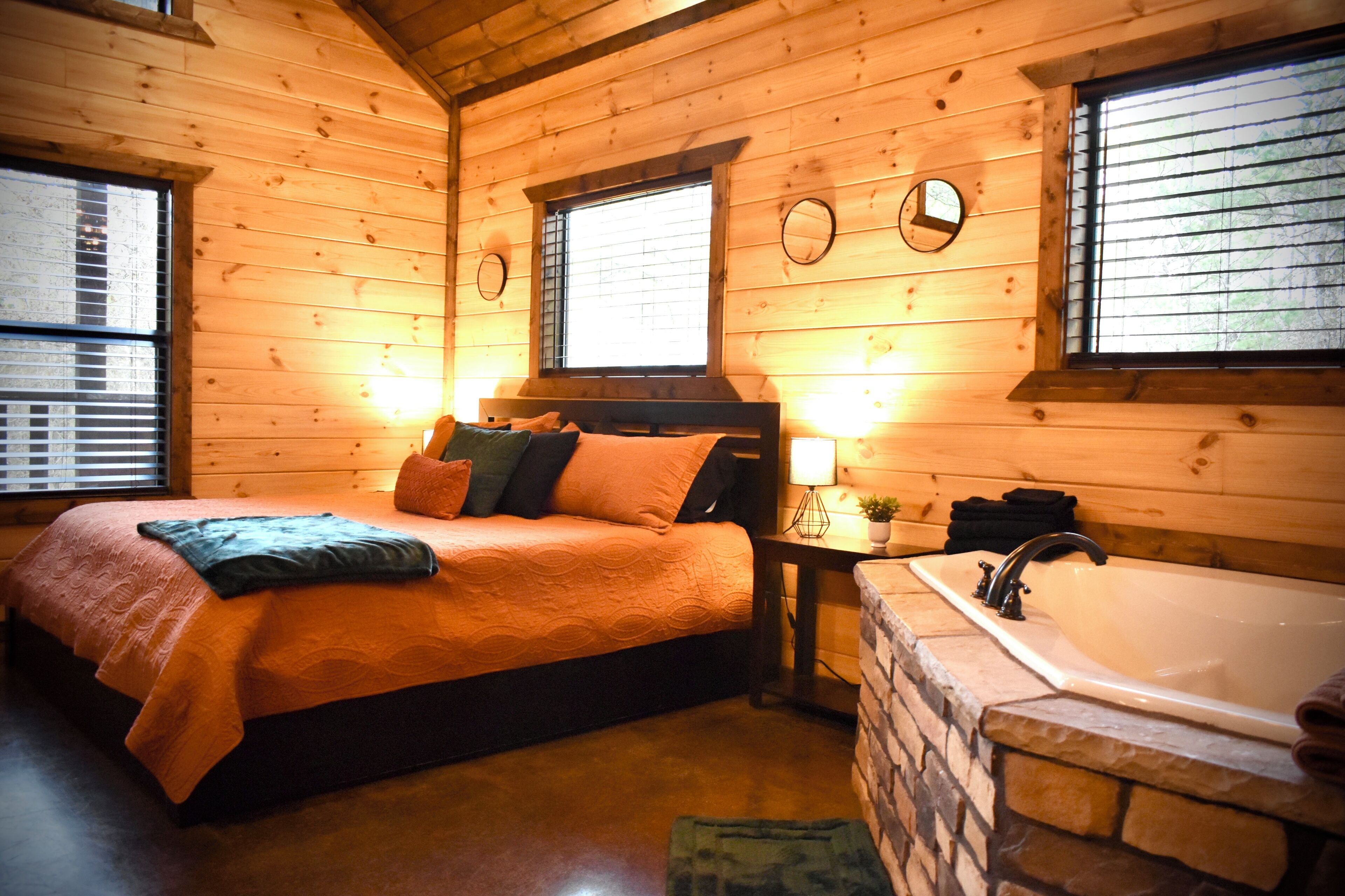 A wooden cabin bedroom with a bed, a nightstand, a lamp, and a stone bathtub.