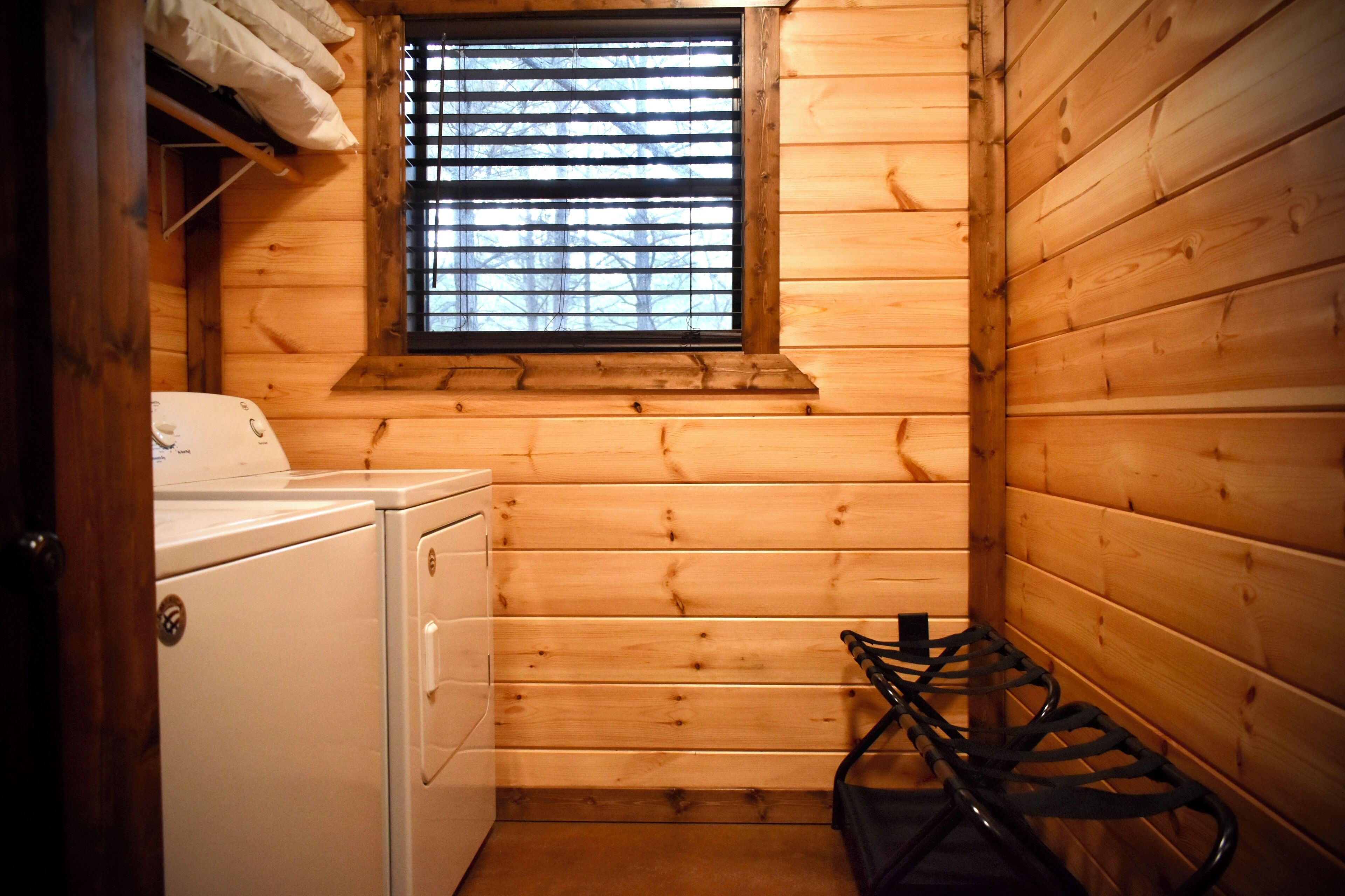 A small, wooden cabin-style room with a washing machine, a window with blinds, and a chair.