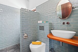 A bathroom with a white toilet, a wooden vanity with a white sink, a mirror, and a shower area with blue tiles.