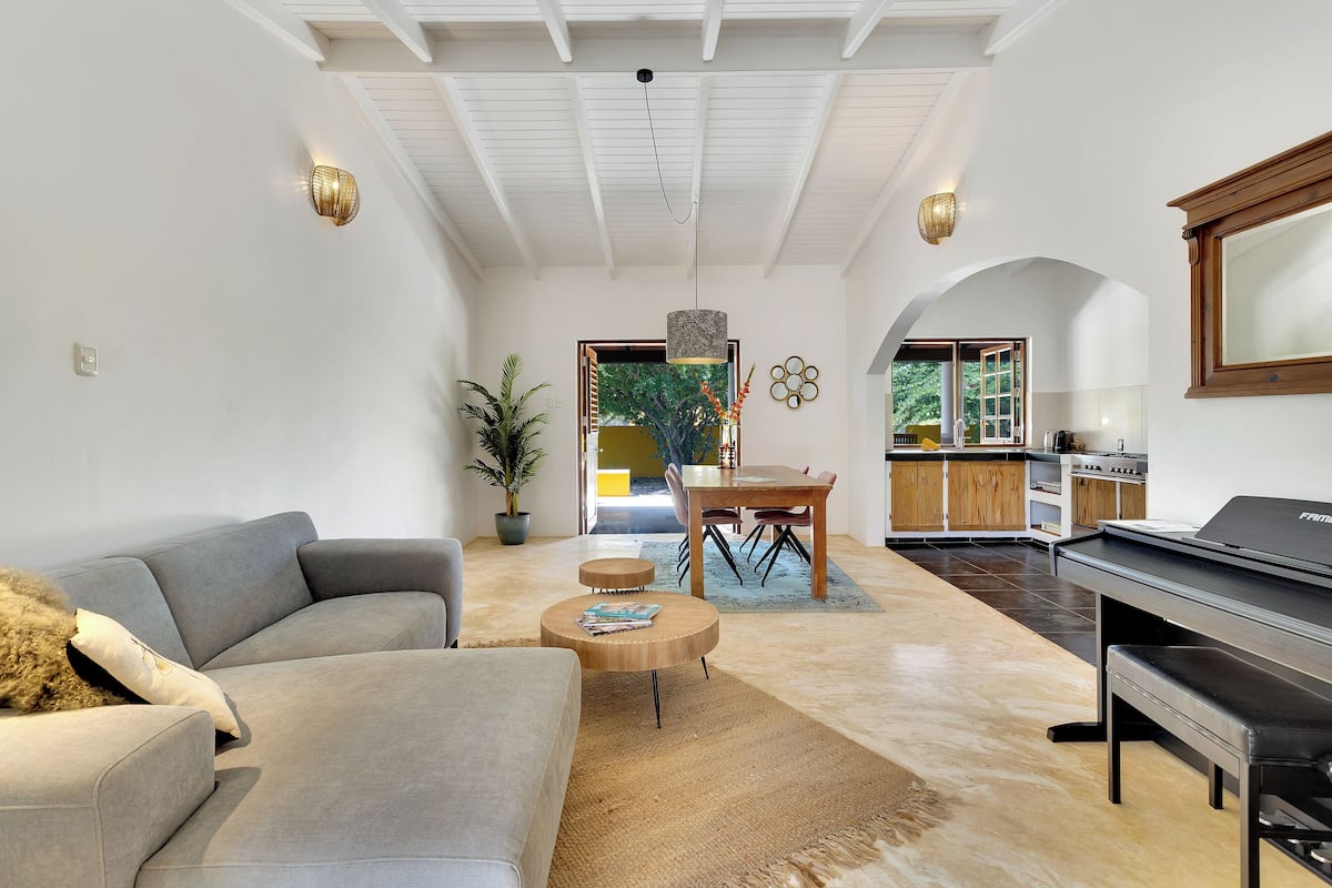 Una sala de estar moderna con un sofá gris, una mesa de centro redonda y un piano. Hay una zona de comedor con una mesa y sillas de madera, y una cocina al fondo.