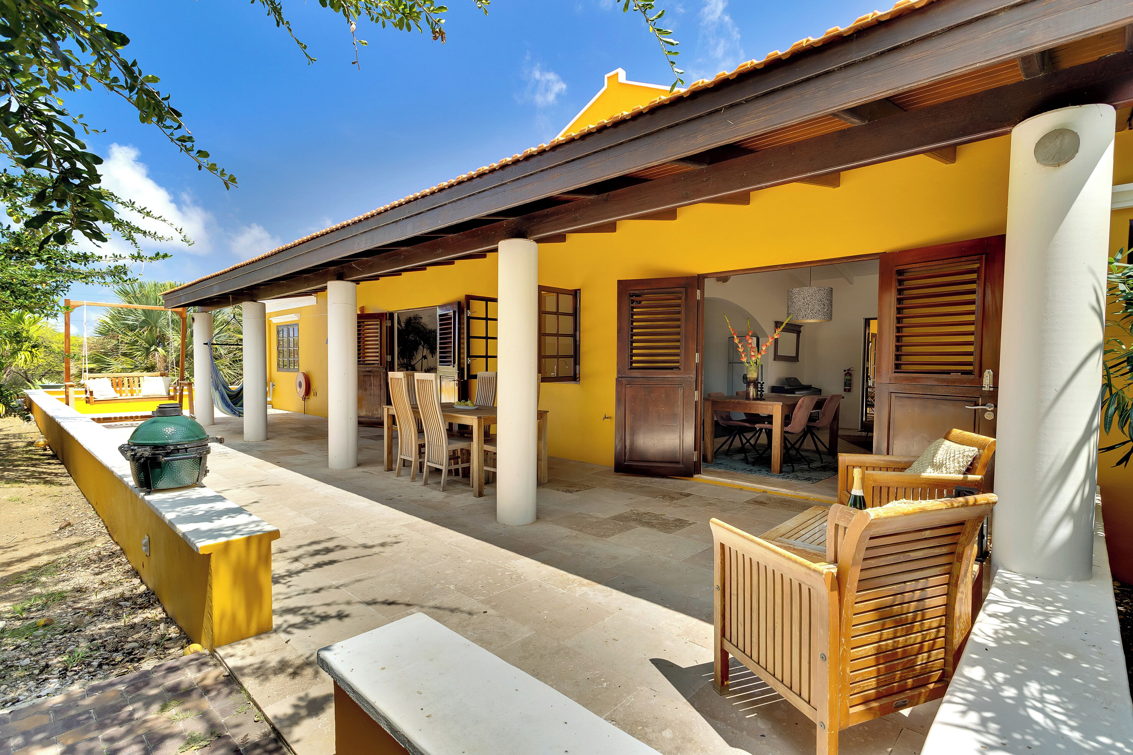 A yellow building with a covered patio, wooden furniture, and a grill.