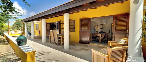 A yellow building with a covered patio, wooden furniture, and a grill.