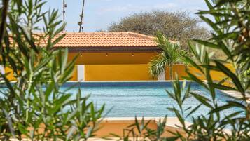 A pool area with a yellow building in the background, surrounded by greenery.