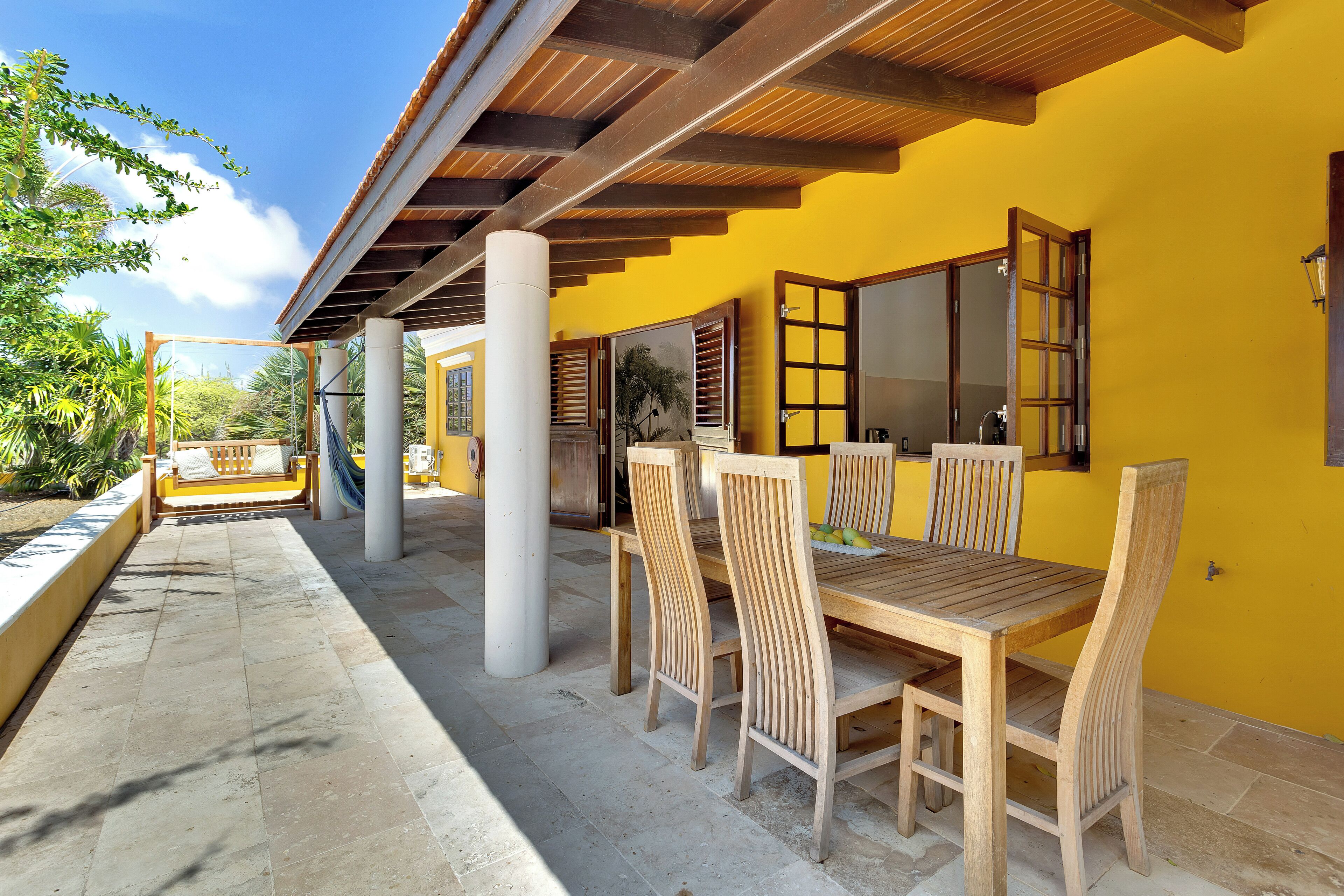 Un patio al aire libre con una mesa y sillas de madera, una pared amarilla y vistas a zonas verdes.