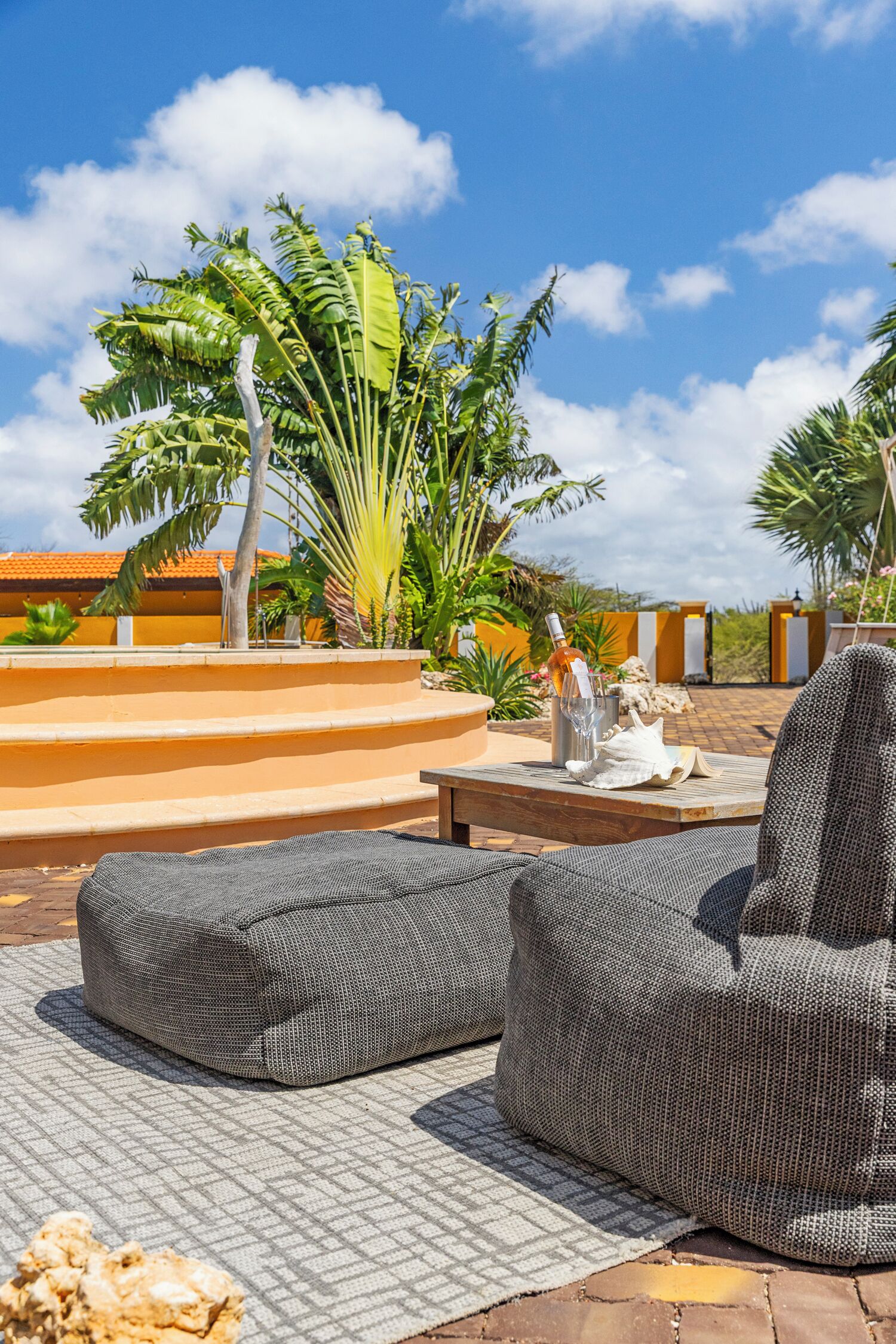 Un espace patio avec deux fauteuils en forme de sac de couchage gris, une petite table et une bouteille de vin. Des marches mènent à une terrasse d’où l’on peut admirer des palmiers et un bâtiment aux murs orange.