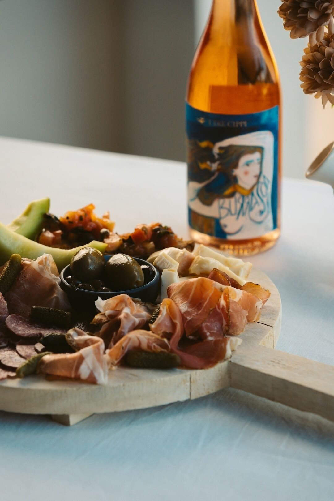 Une planche de charcuterie avec différentes sortes de viandes et d’olives, une bouteille de vin et un élément décoratif.