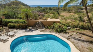 A swimming pool with a stone sculpture, surrounded by a landscaped garden and a view of the ocean. - Oceanview Condo With Pool Near Playa Prieta 2BR - Vida de la Paz 4 (Tempate)