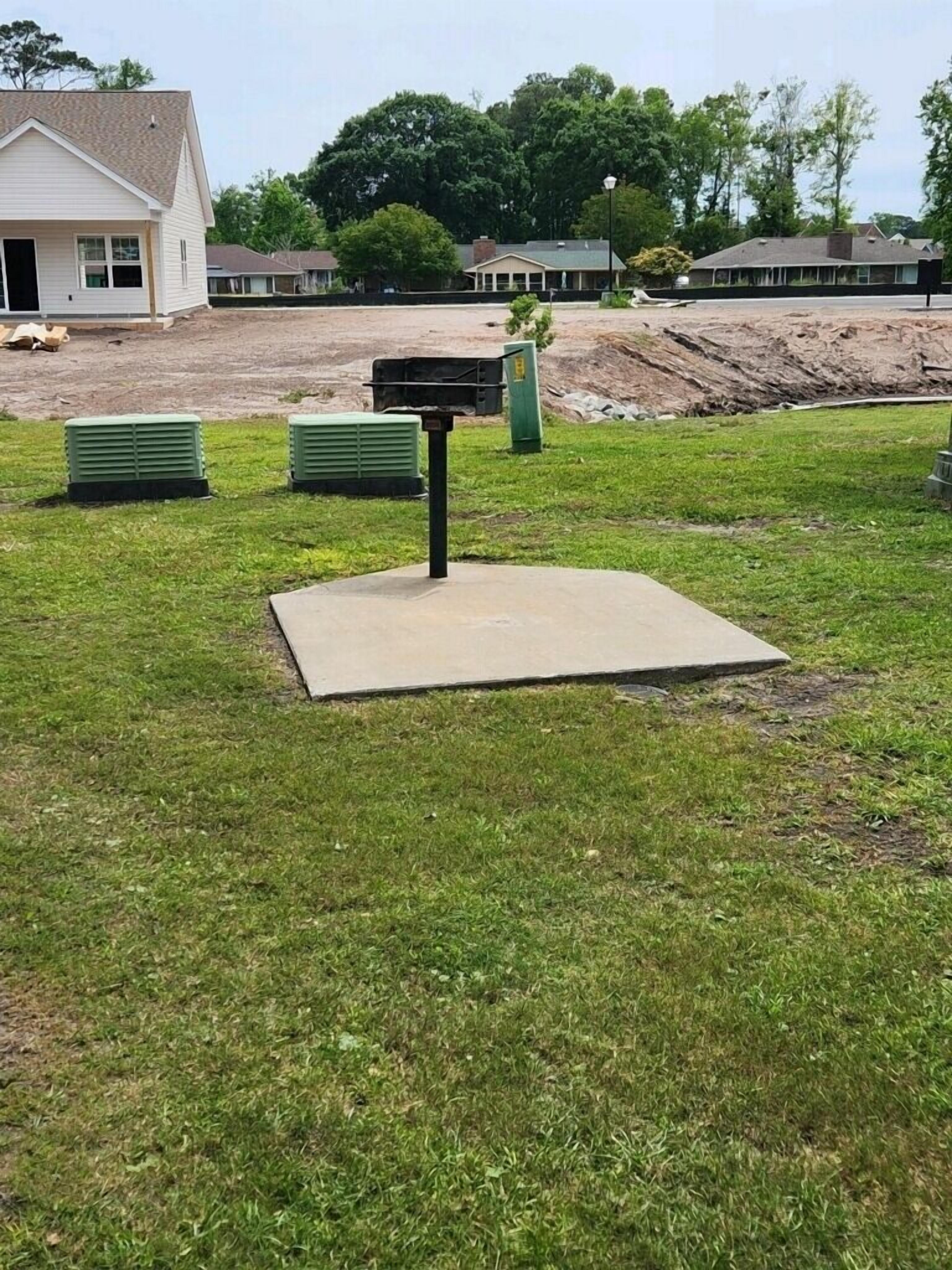 Une zone herbeuse avec une dalle de béton, un coffret électrique vert et un bâtiment en arrière-plan.