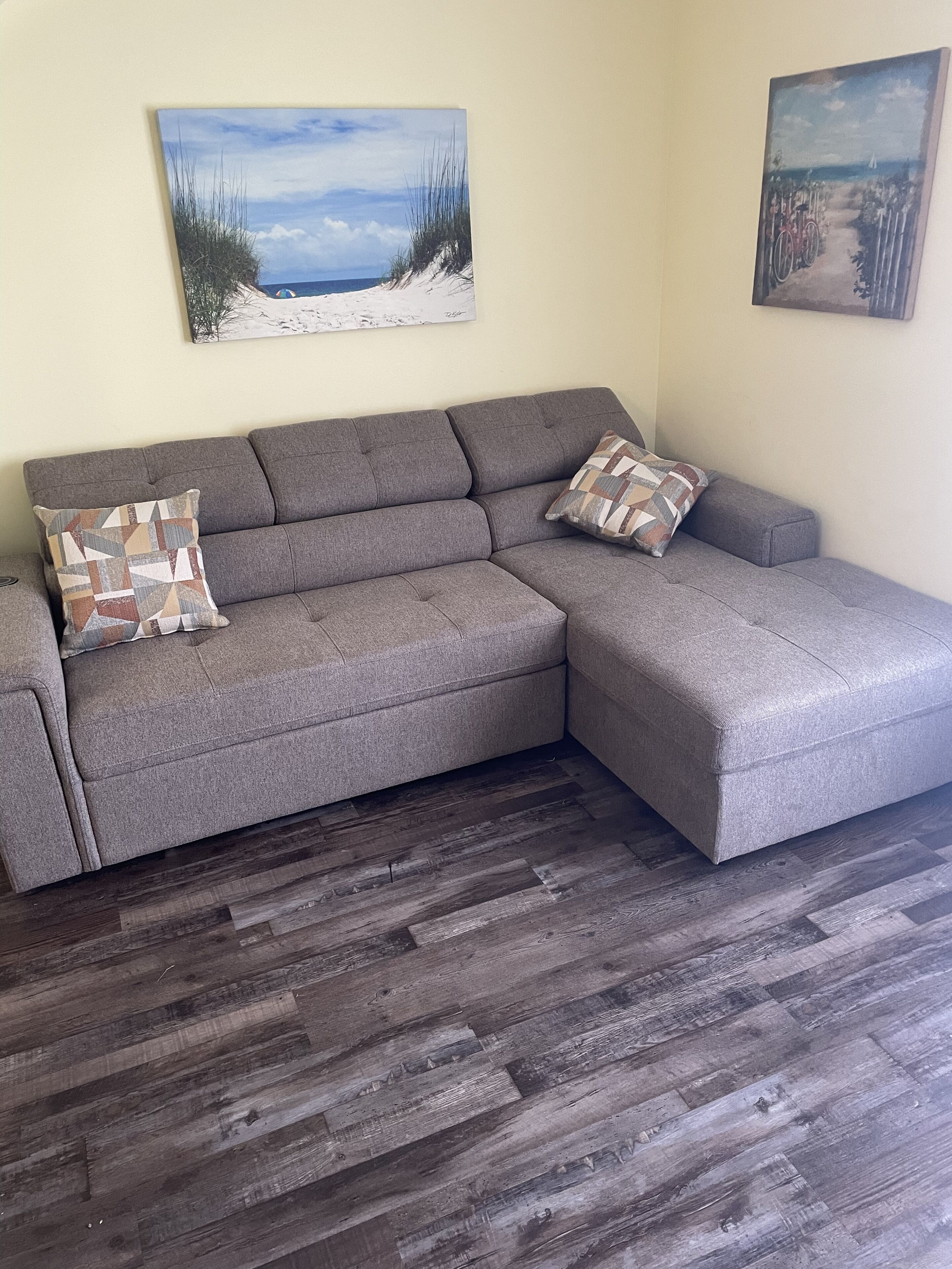 Un canapé d’angle gris avec deux coussins à motifs, un plancher en bois et deux tableaux accrochés au mur.