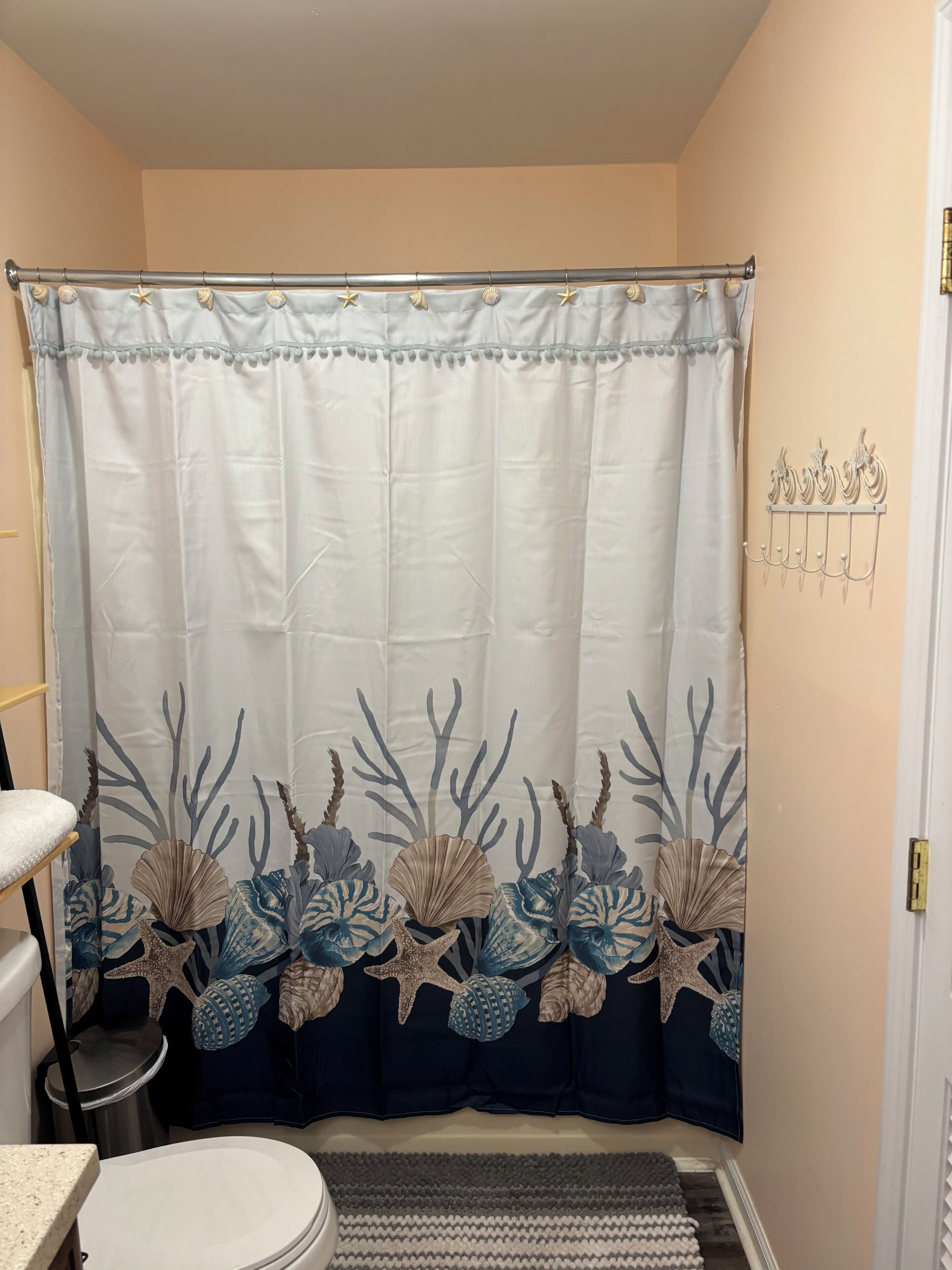 Un baño con cortina de ducha de diseño marino, un inodoro, un pequeño cubo de basura y un perchero de pared.