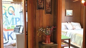 Une pièce chaleureuse aux murs en bois, un lit recouvert d’une literie blanche, une petite table ornée d’un vase de fleurs et une porte sur laquelle est accrochée une enseigne colorée.