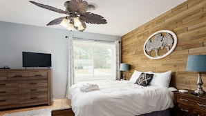 Ein Schlafzimmer mit Bett, Holz-Kopfteil, Deckenventilator, Fernseher, Holzkommode und einem Fenster mit Vorhängen.
