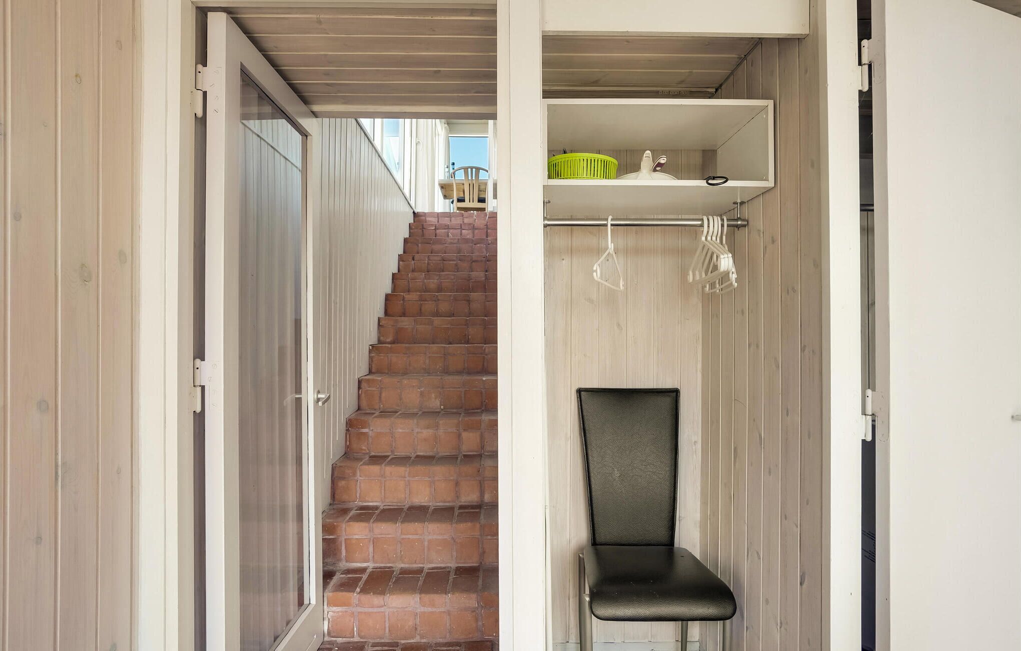 A narrow hallway with a staircase leading to a balcony. A chair is positioned against the wall, and a shelf with a green basket is visible.