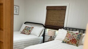 Una camera con due letti singoli, ognuno con testiera e cuscino. Si vede una finestra con tenda, una lampada a parete e un quadro incorniciato.