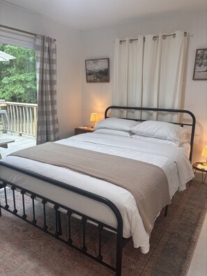 Un dormitorio con una cama, una mesita de noche con lámpara, una ventana con cortinas y un cuadro en la pared.