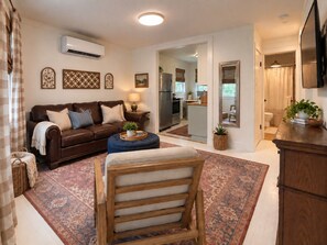 Una sala de estar con un sofá, un reposapiés y una silla de madera. Se ve una cocina a través de un vano abierto.