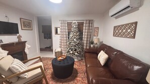 Een woonkamer met een bruine leren bank, een zwart voetenbankje, een houten stoel, een televisie op een standaard, een kerstboom en een kamerkleed met een patroon.