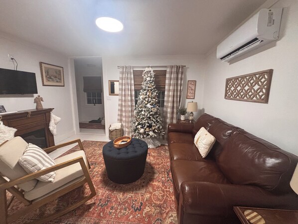 Un salon avec un canapé en cuir marron, un pouf noir, une chaise en bois, un téléviseur posé sur un meuble, un sapin de Noël et un tapis à motifs.