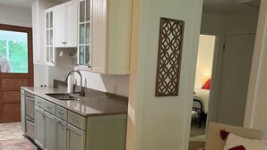 Een knus woongedeelte met een kitchenette, een eethoek met een stoel en tafel, en uitzicht door een geopende deur.