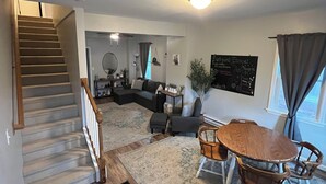 Een woonkamer met een trap, een eethoek met een ronde tafel en stoelen, een bank en een aan de muur bevestigd bord.