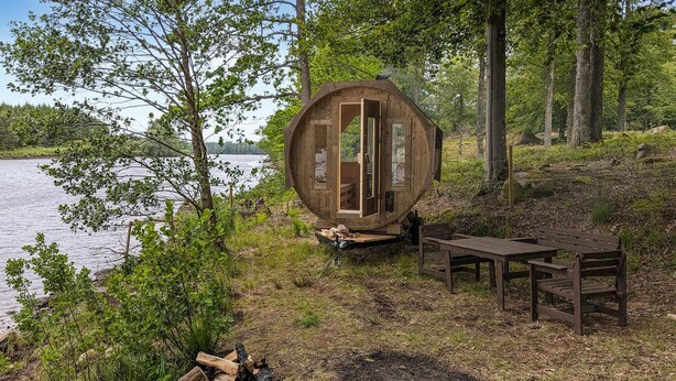 Une cabane en bois dotée d’une grande porte circulaire, d’une table et de bancs, située au bord d’un lac entouré d’arbres.