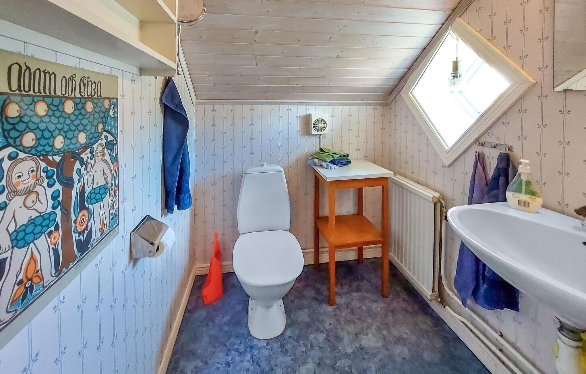 Ein kleines, rustikales Badezimmer mit Toilette, Holzstuhl und einem kleinen Tisch. Es gibt eine Heizung, ein Fenster mit Blick und eine Wand mit einem farbenfrohen Wandgemälde.