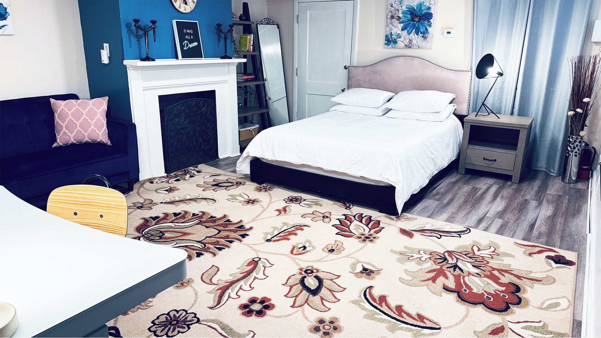 Un dormitorio con cama, mesita de noche, cómoda, silla, escritorio, chimenea y una pared con reloj y obras de arte.