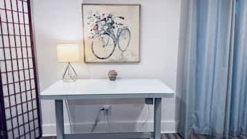 Une table blanche avec une lampe et une petite plante en pot, un tableau encadré représentant un vélo et une prise de courant.
