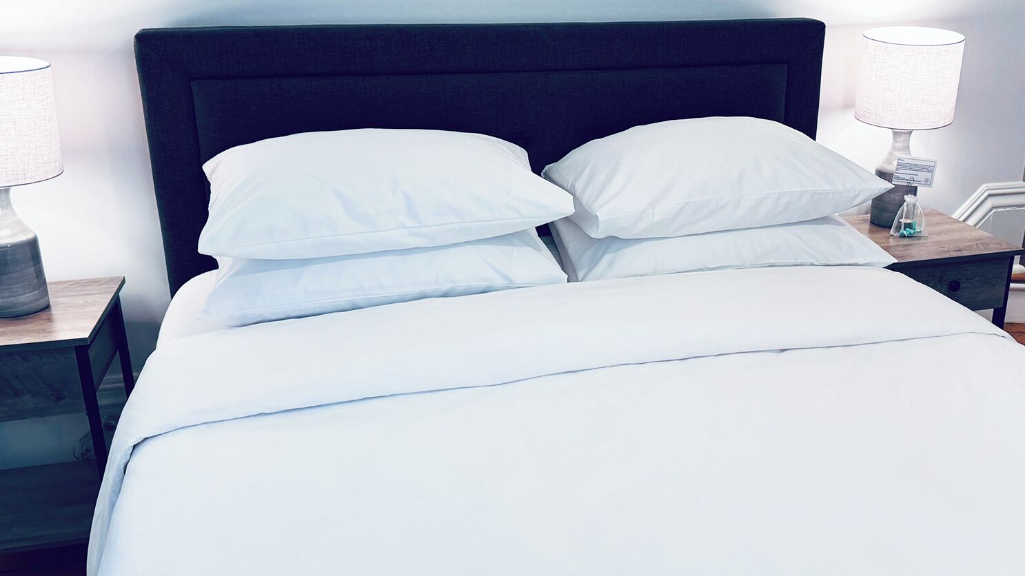 Una cama bien tendida con sábanas y almohadas blancas, un cabecero oscuro y lámparas de noche sobre mesitas de madera.