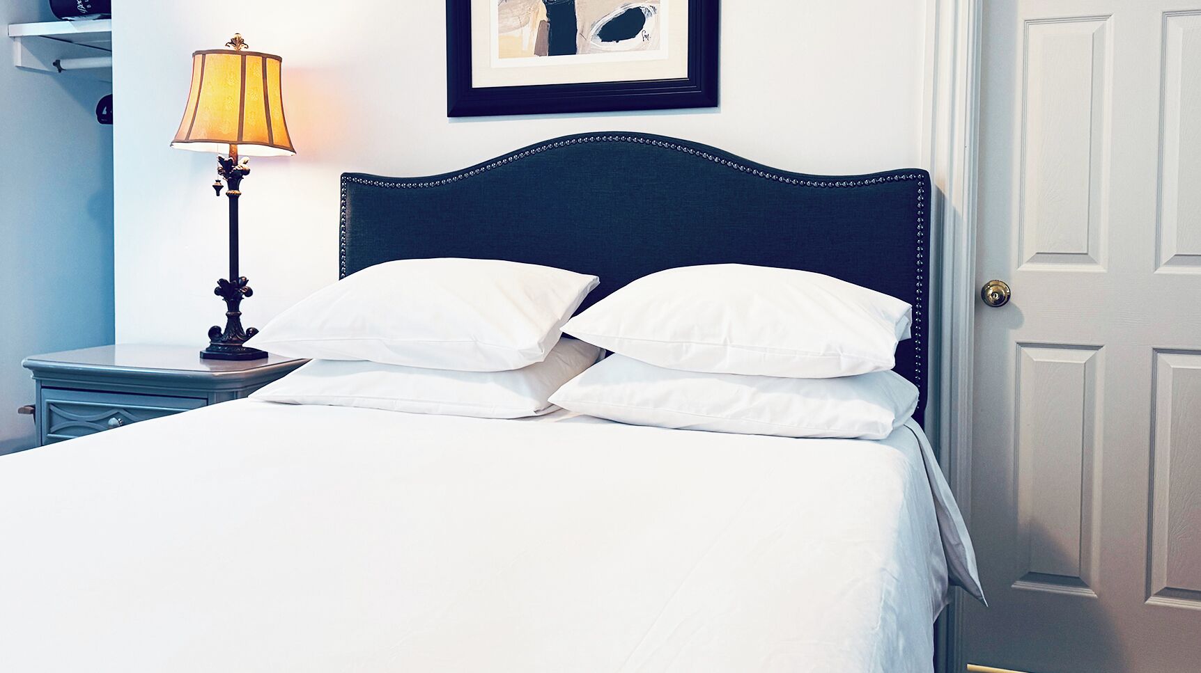 Una cama bien hecha con ropa blanca y un cabecero oscuro, una mesita de noche con una lámpara y un cuadro en la pared.
