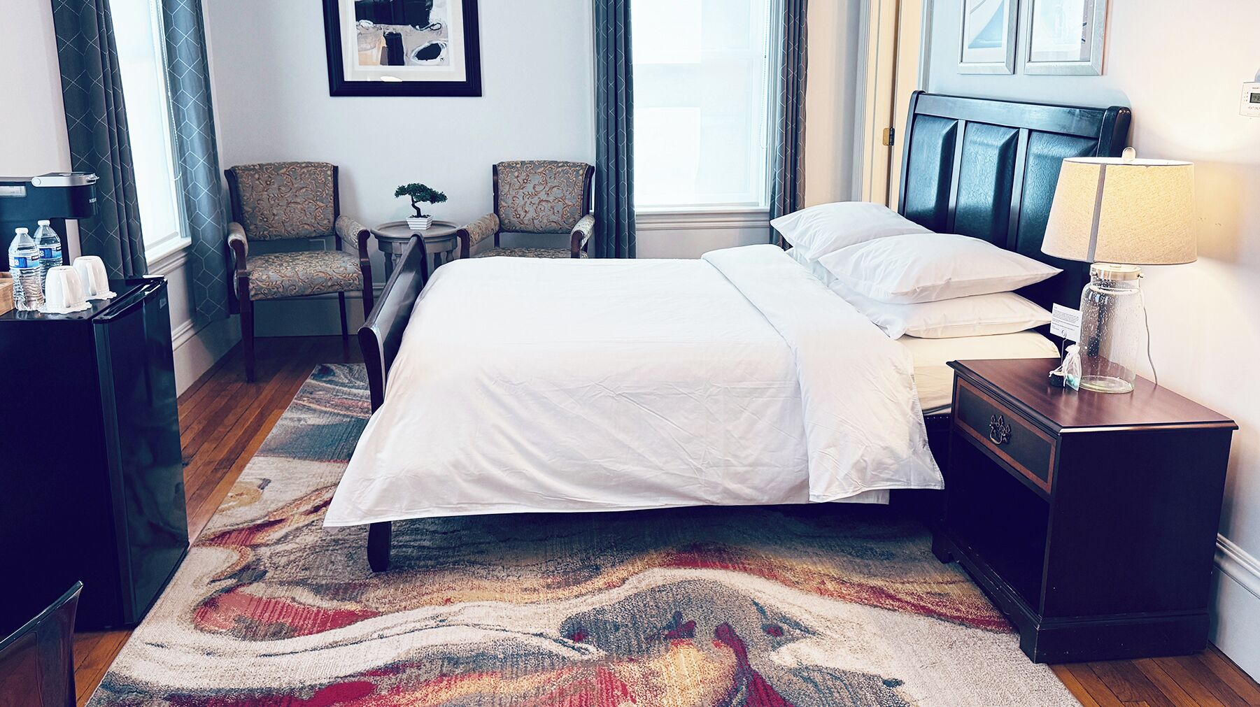 A hotel room with a bed, two chairs, a small table with a bonsai tree, a nightstand with a lamp, and a water dispenser.