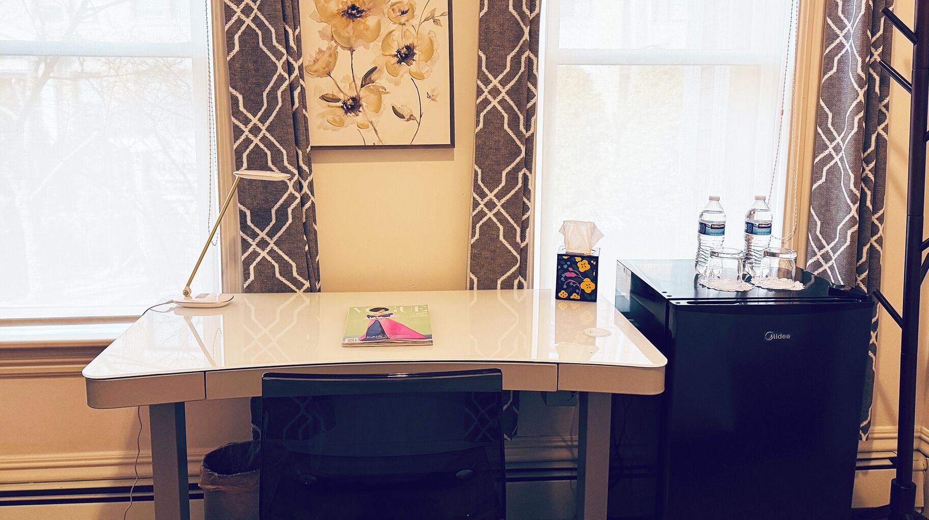 A hotel room with a white desk, a framed floral artwork on the wall, a small table with a tissue box and water bottles, and a black minibar.