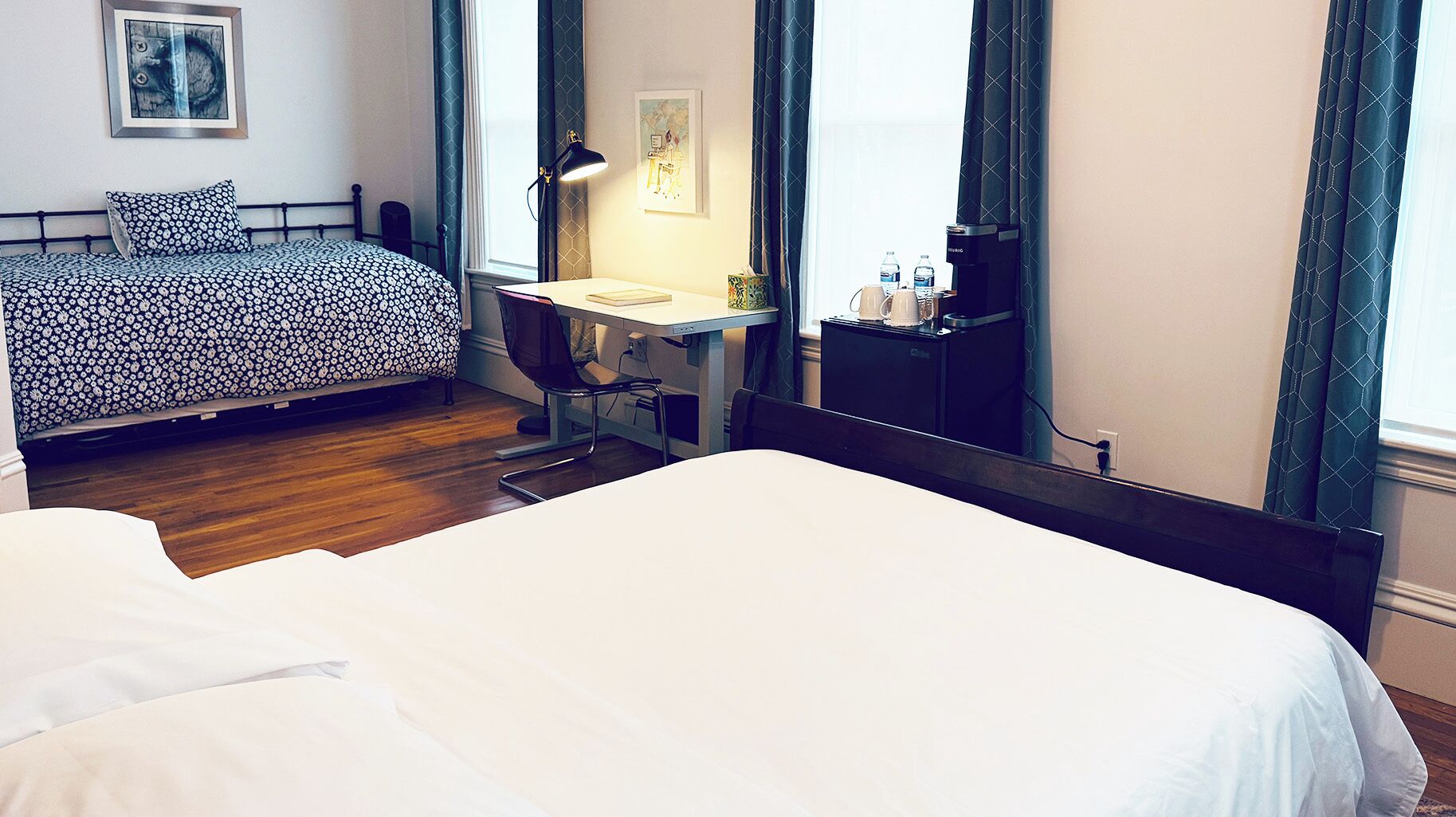 A hotel room with a large bed, a desk with a chair, a desk lamp, a small table with a kettle, and a window with curtains.