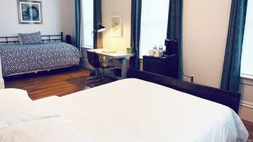 A hotel room with a large bed, a desk with a chair, a desk lamp, a small table with a kettle, and a window with curtains.